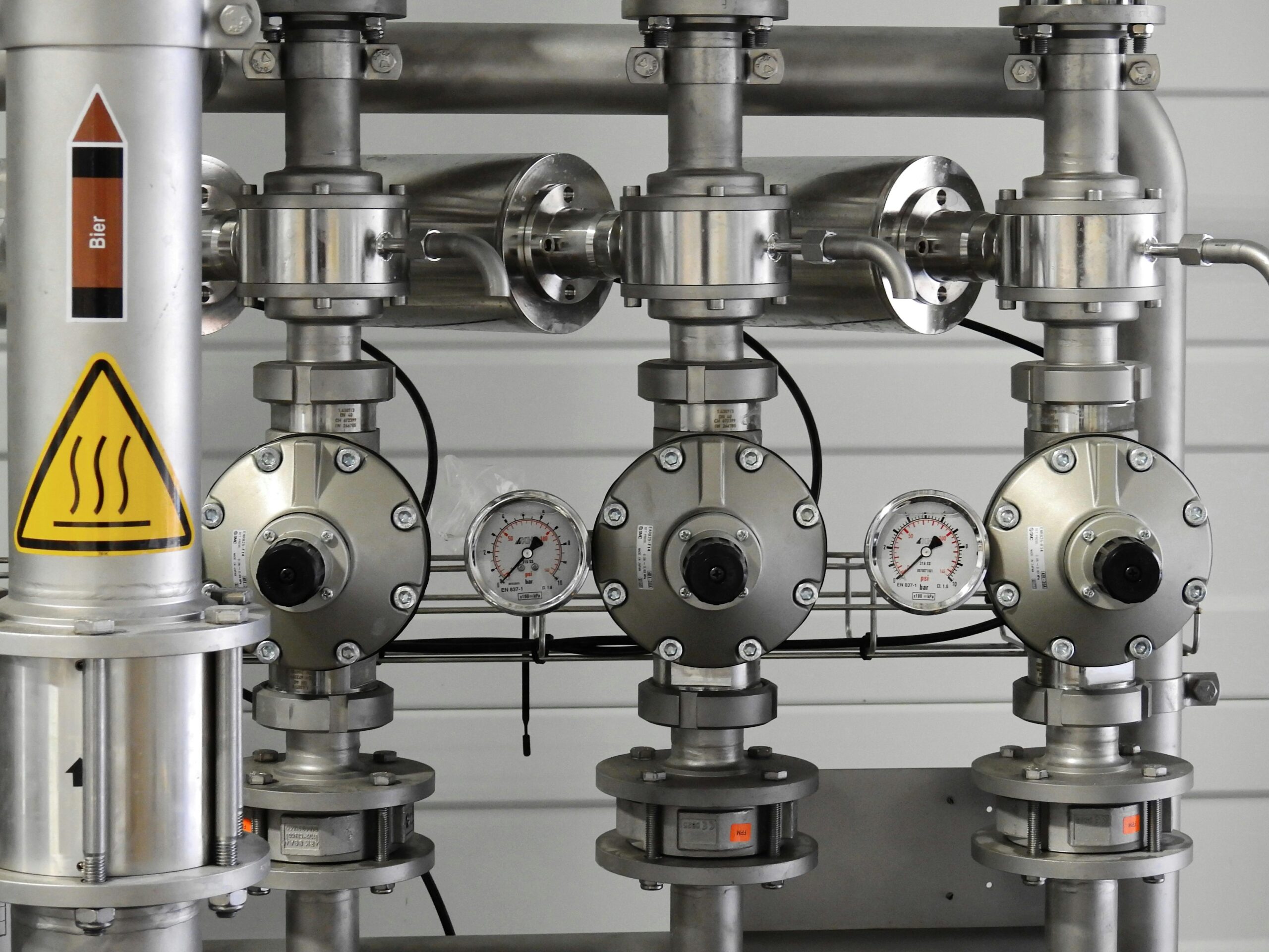 A close-up view of an industrial piping system with stainless steel pipes, pressure gauges, and control valves. A warning sign with a heat symbol and the word "Bier" indicates the system is related to beer production or brewing. The gauges display pressure readings, highlighting the controlled process of fluid transfer.